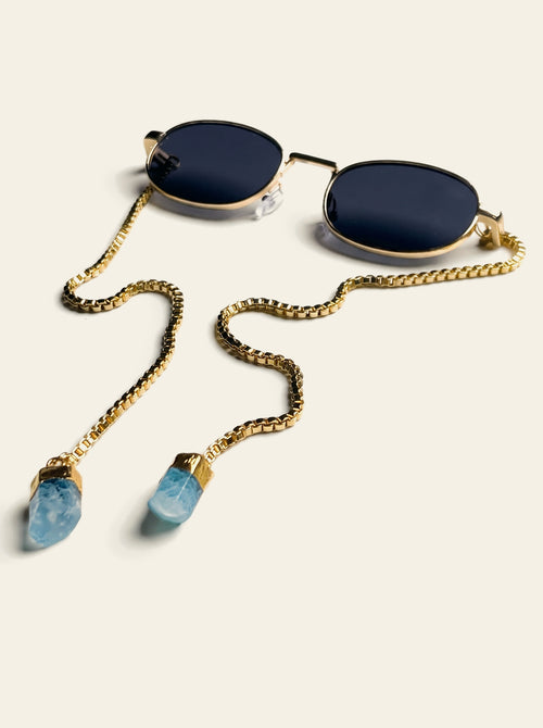 The Amalfi - gold oval sunglasses with solid navy-black lenses and a gold chain ending in two raw light-blue quartz stones, three-quarter view