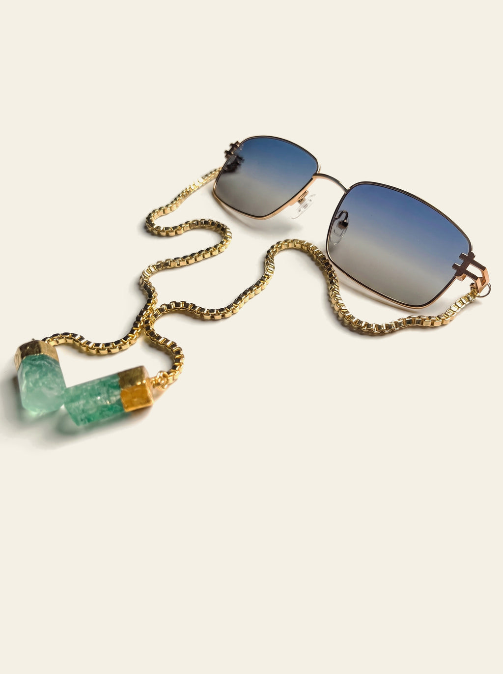 The Ravello - thin gold square frame sunglasses with blue-grey gradient lenses and a gold box chain ending in two raw green quartz stones, three-quarter view