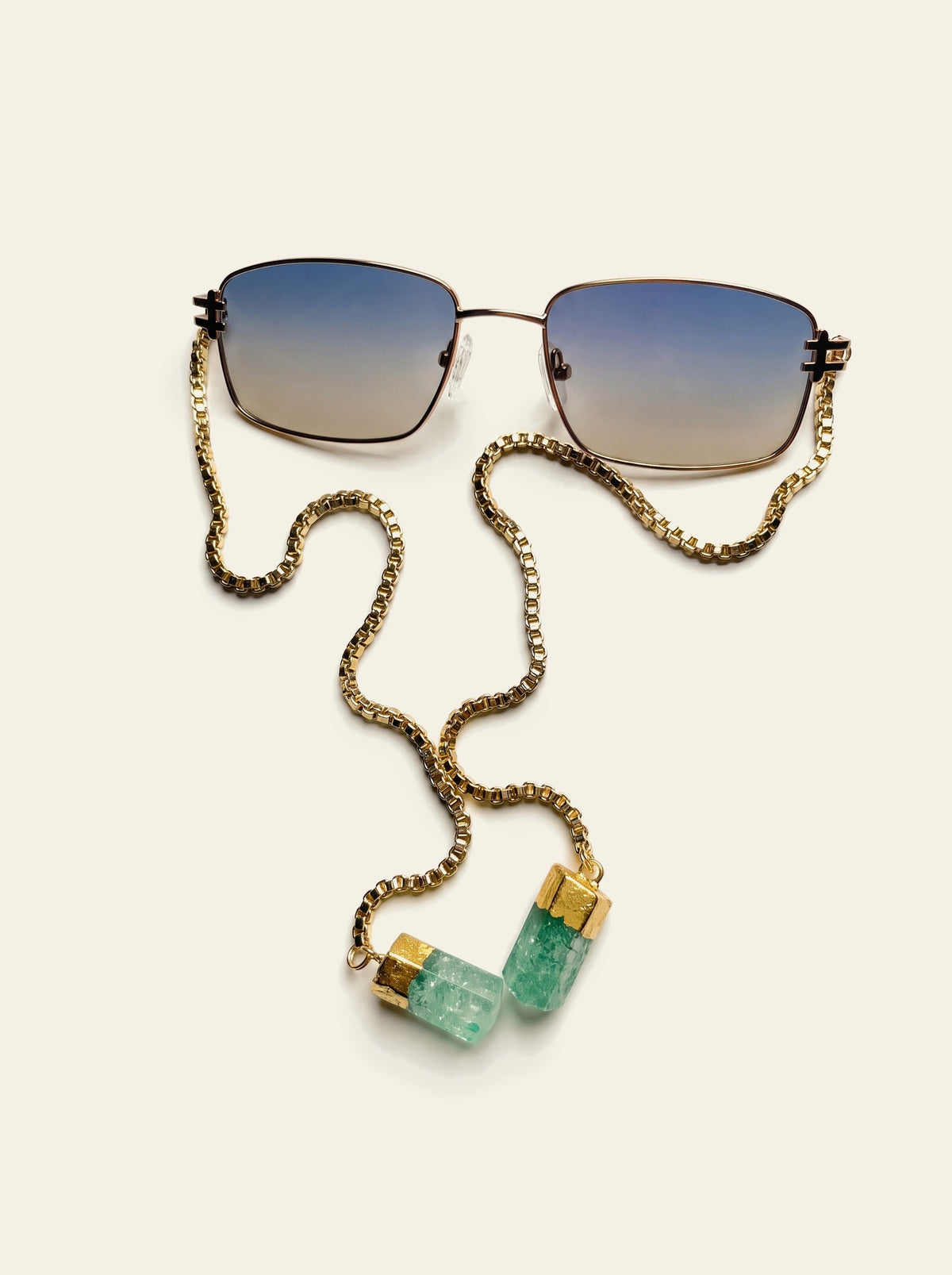 The Ravello - top view of gold square frame sunglasses with blue-grey gradient lenses, gold chain draped to two green quartz stones