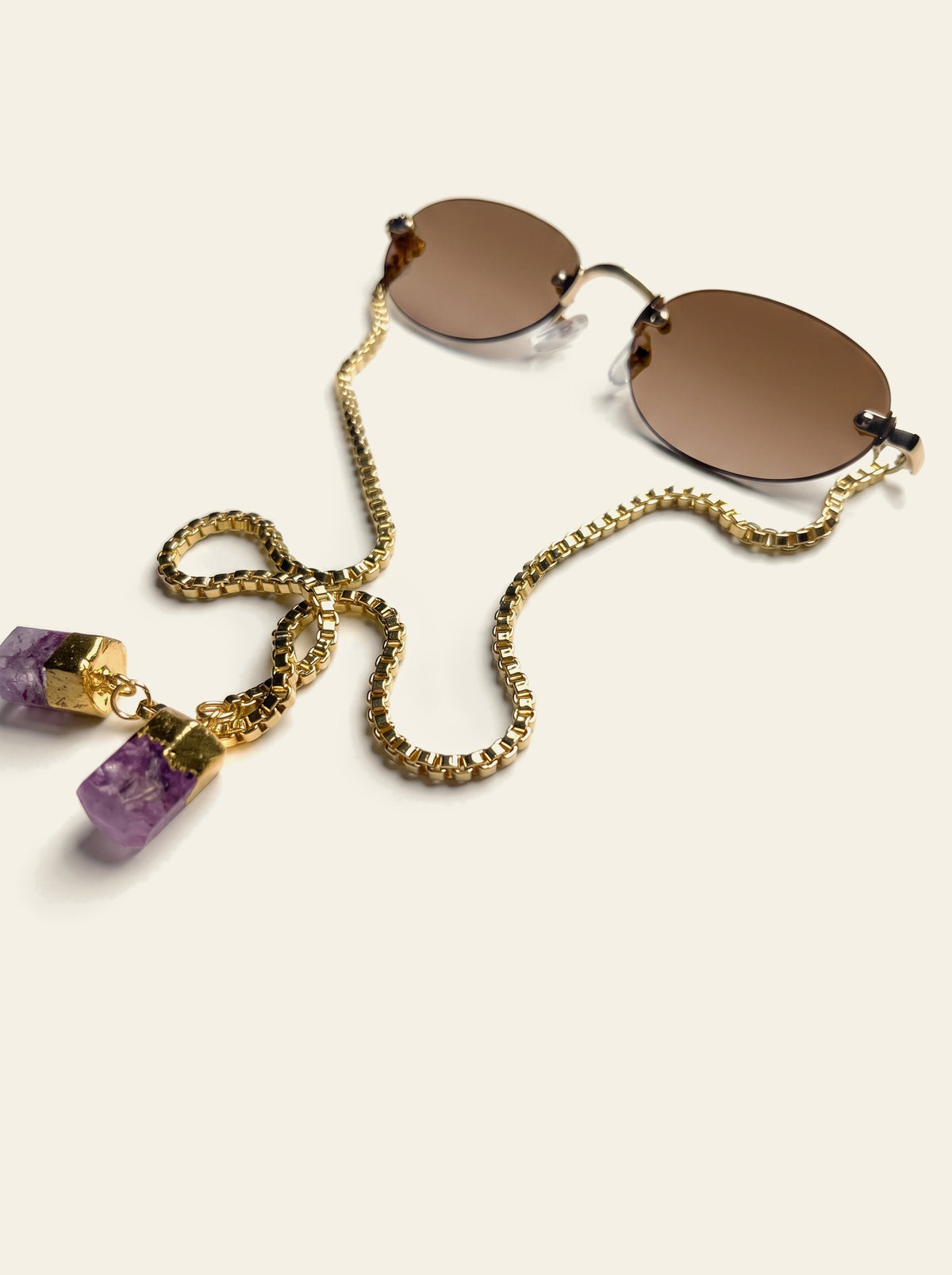 The Ischia - rimless oval sunglasses with amber-brown lenses, gold bridge and end pieces, and a gold box chain ending in two raw purple quartz stones, three-quarter view