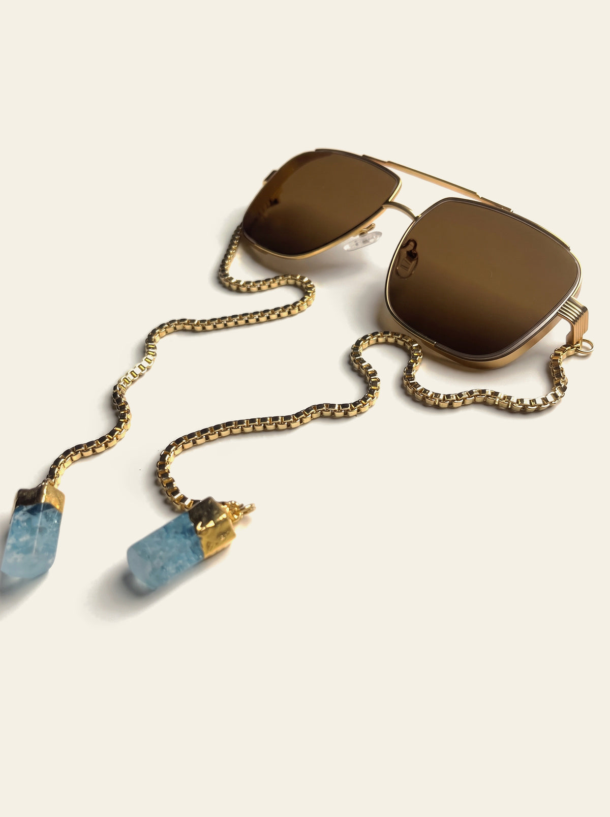 The Como - gold double-bridge navigator sunglasses with brown lenses, three-quarter view, gold box chain draping to two raw blue quartz stones