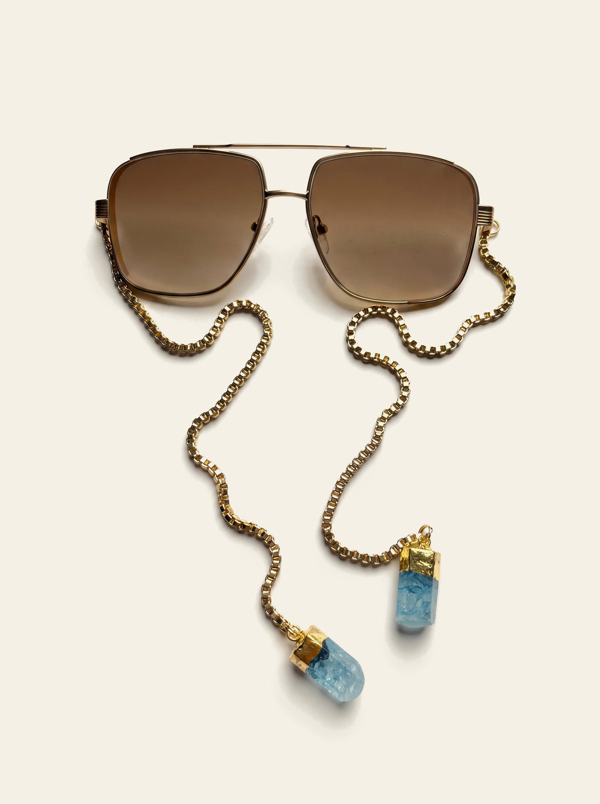 The Como - top view of gold double-bridge navigator frame with brown lenses, gold chain and two pale blue quartz stones laid out below