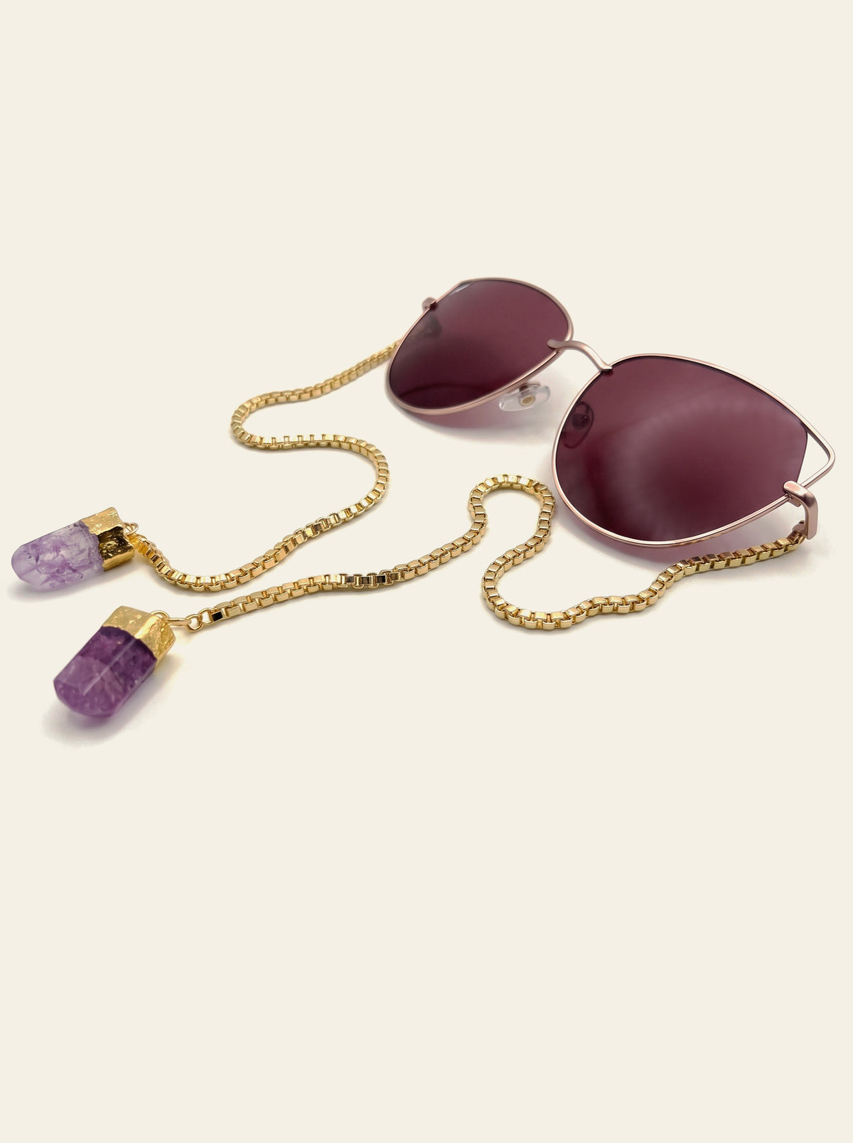 The Taormina - three-quarter view of gold thin-rim cat-eye sunglasses with solid burgundy lenses and a gold chain ending in two raw purple quartz stones