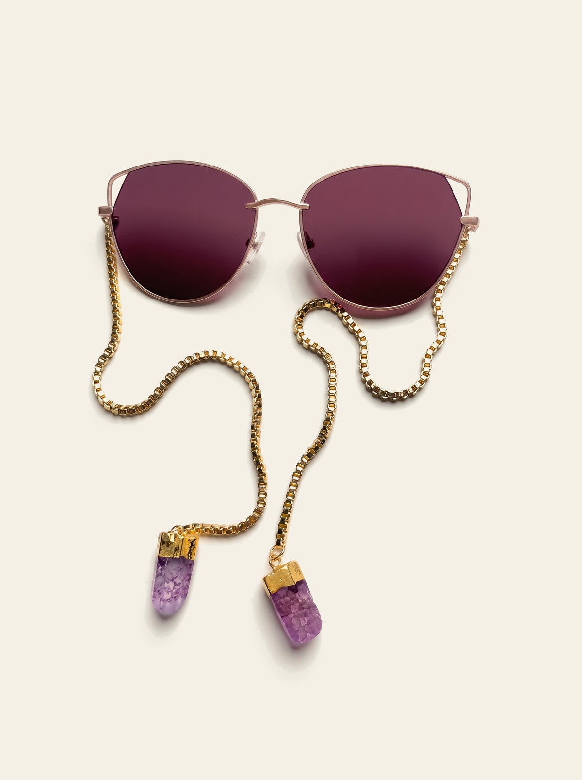 The Taormina - top view of gold cat-eye sunglasses with burgundy lenses, the gold box chain laid out to two purple quartz stones in gold caps