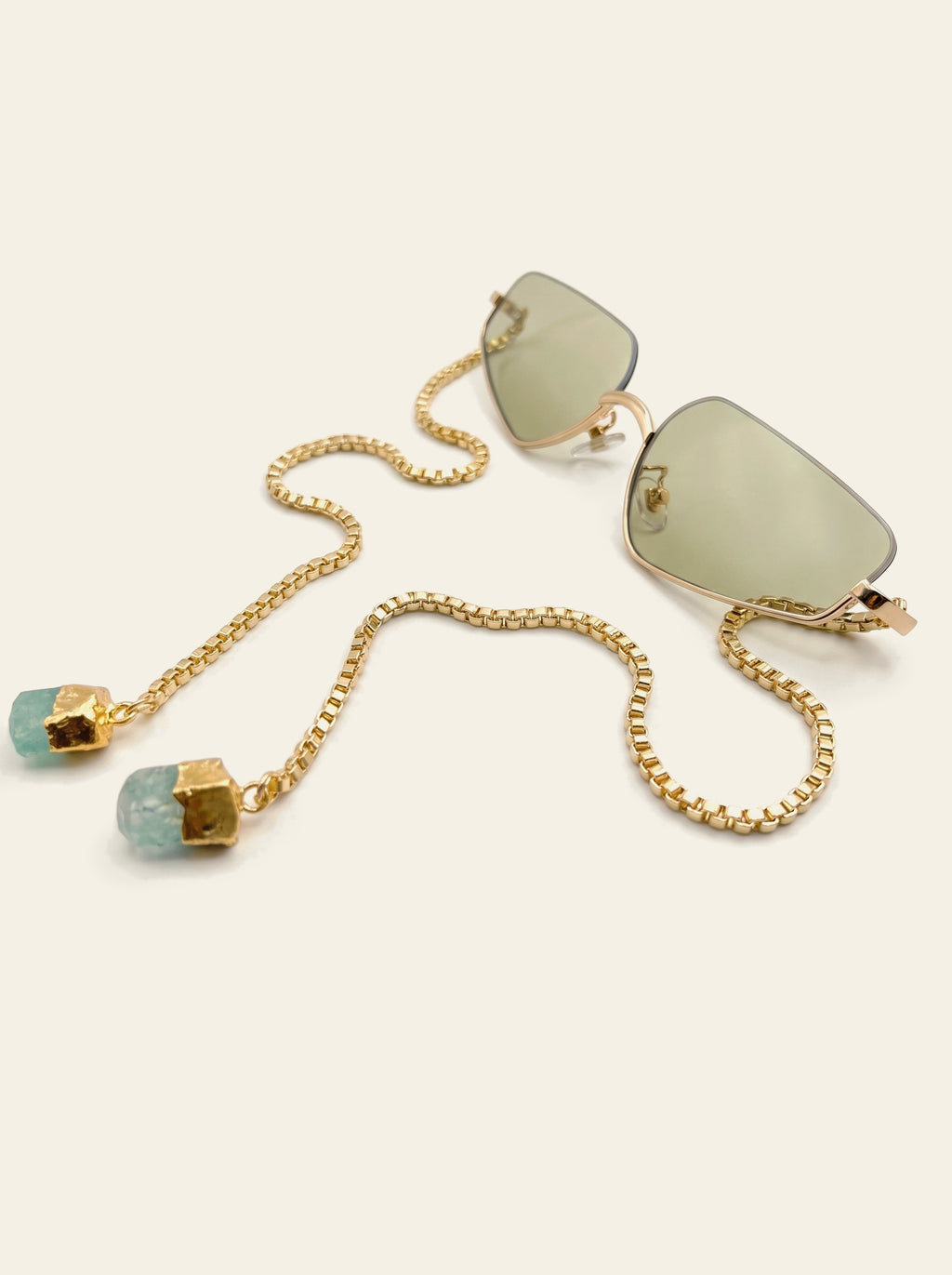 The Antibes - rimless gold rectangle sunglasses with pale green lenses, three-quarter view, gold box chain and two raw green quartz stones resting beside the frame