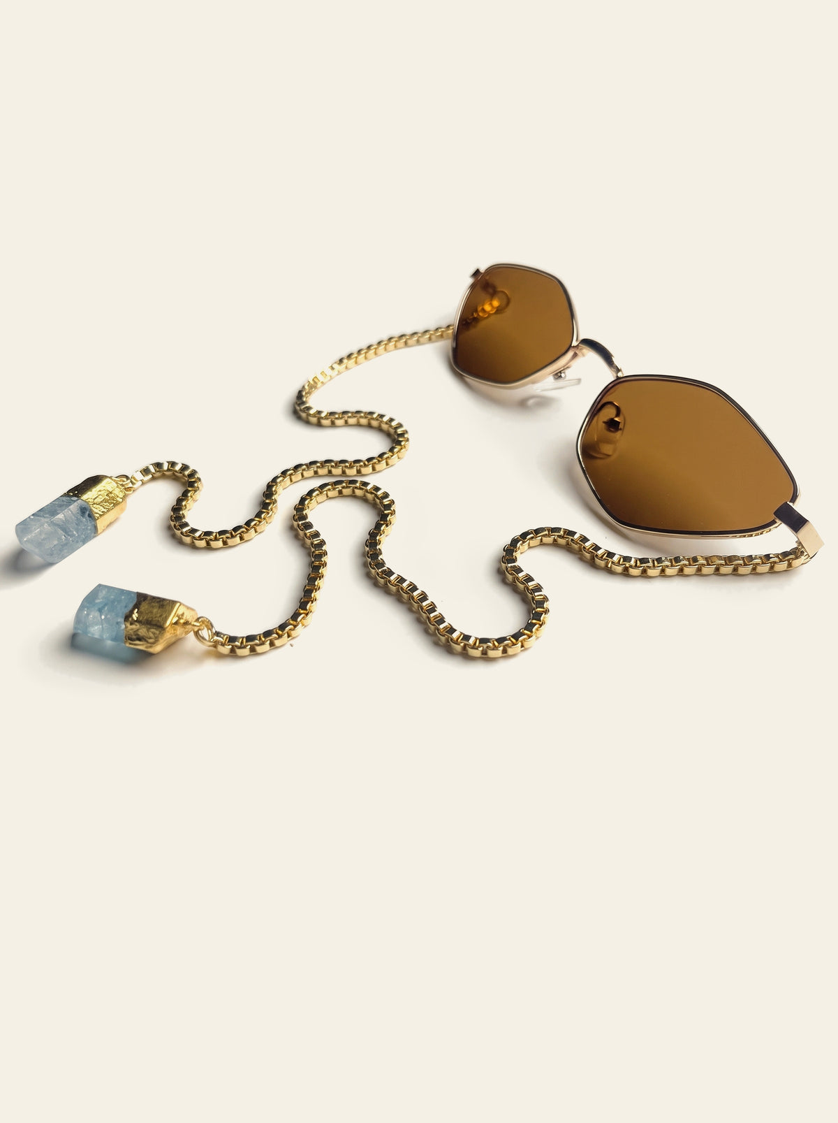 The Cassis - thin gold hexagon sunglasses with solid brown lenses, seen from the side with the gold chain and two pale blue quartz stones