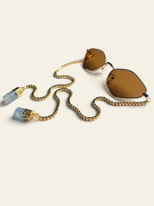The Cassis - thin gold hexagon sunglasses with solid brown lenses, seen from the side with the gold chain and two pale blue quartz stones