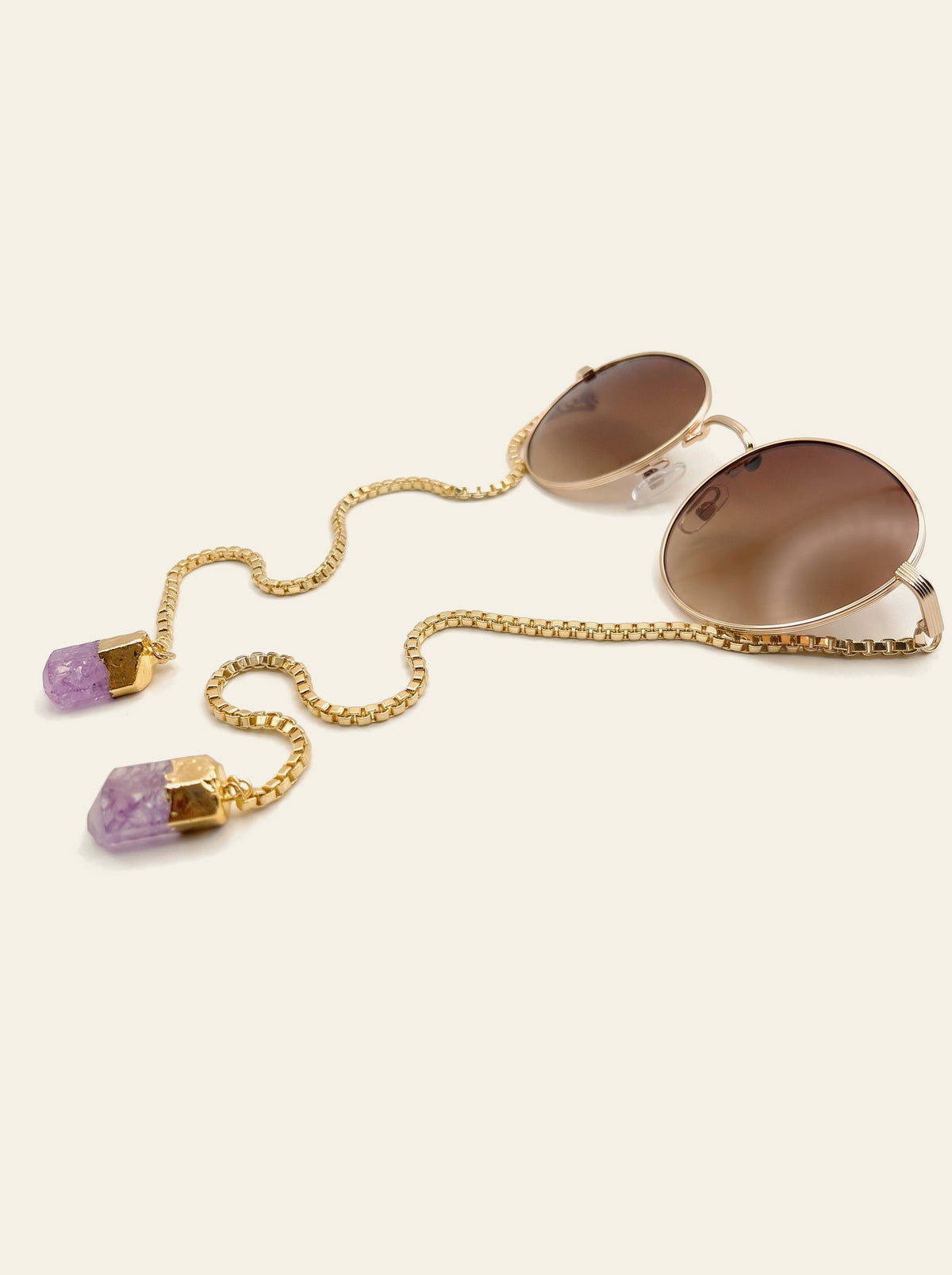 The Procida - three-quarter view of round gold wire-rim sunglasses with solid brown lenses and a gold box chain ending in two raw purple quartz stones