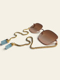 The Menton - square gold-finished sunglasses with brown gradient lenses, three-quarter view, with gold chain and two raw blue quartz stones