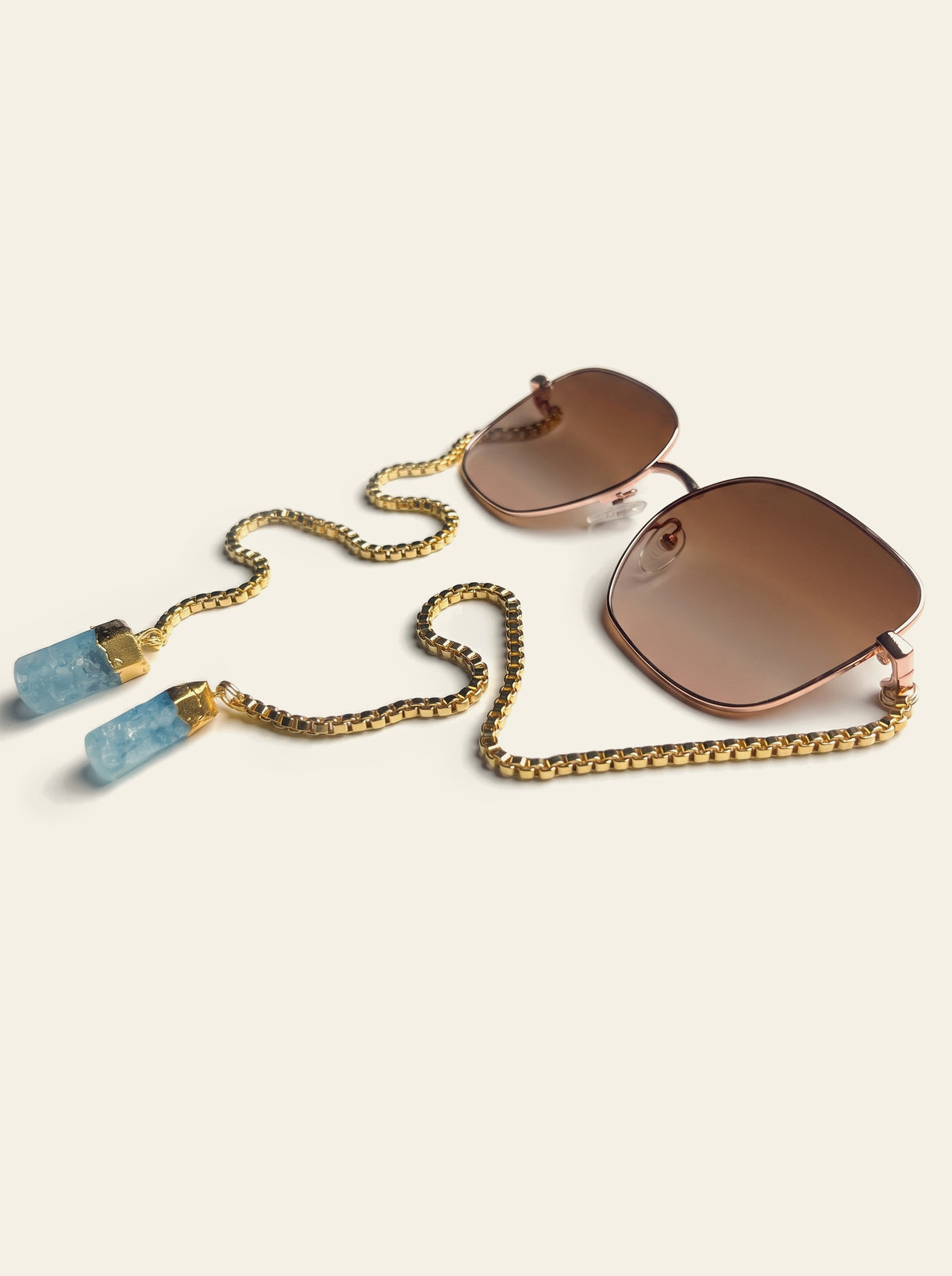 The Menton - square gold-finished sunglasses with brown gradient lenses, three-quarter view, with gold chain and two raw blue quartz stones