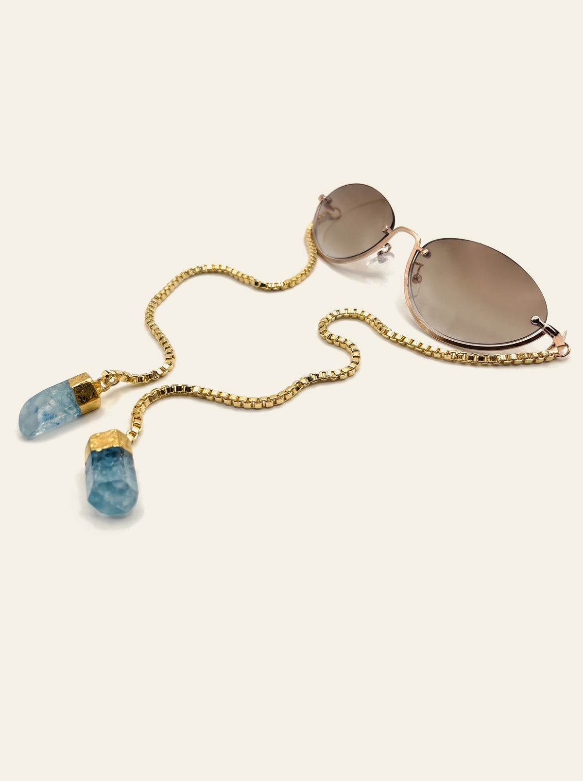 The Rapallo - rimless oval sunglasses with brown gradient lenses, gold wire frame and gold chain ending in two raw blue quartz stones, three-quarter view