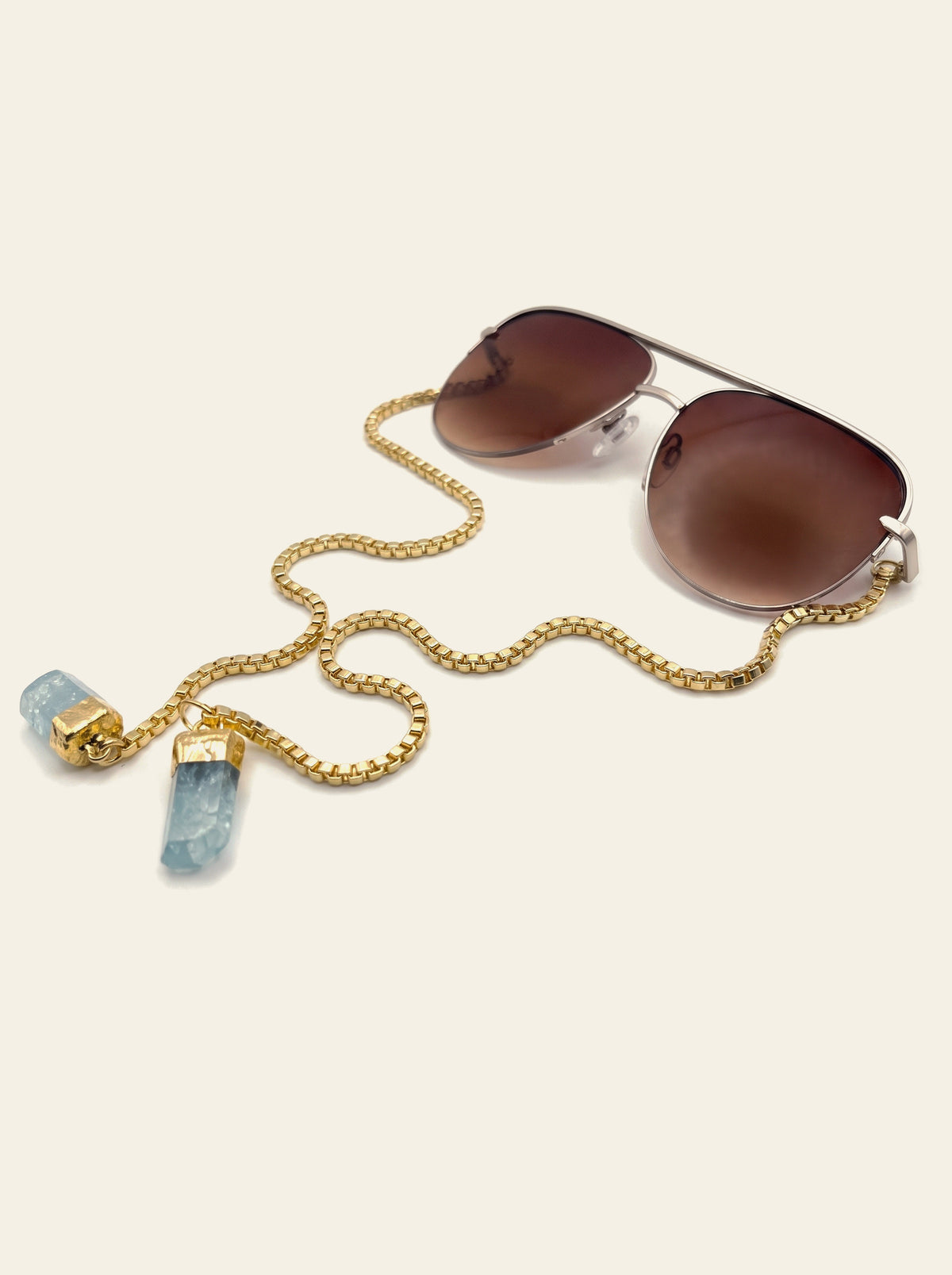 The Camogli - gold semi-rimless aviator sunglasses with brown gradient lenses, seen from three-quarter view, gold chain trailing to two raw blue quartz stones