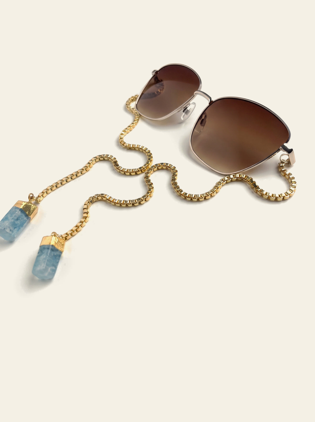 The Biarritz - gold semi-rimless butterfly sunglasses with brown gradient lenses and a gold chain ending in two raw blue quartz stones, three-quarter view