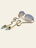 The Stresa - rimless oval sunglasses with gold bridge, blue-grey gradient lenses and a gold box chain ending in two raw pale blue quartz stones, three-quarter view