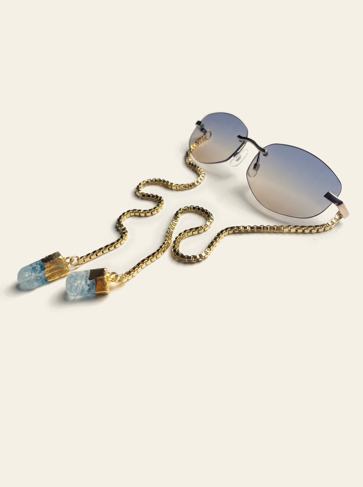 The Stresa - rimless oval sunglasses with gold bridge, blue-grey gradient lenses and a gold box chain ending in two raw pale blue quartz stones, three-quarter view