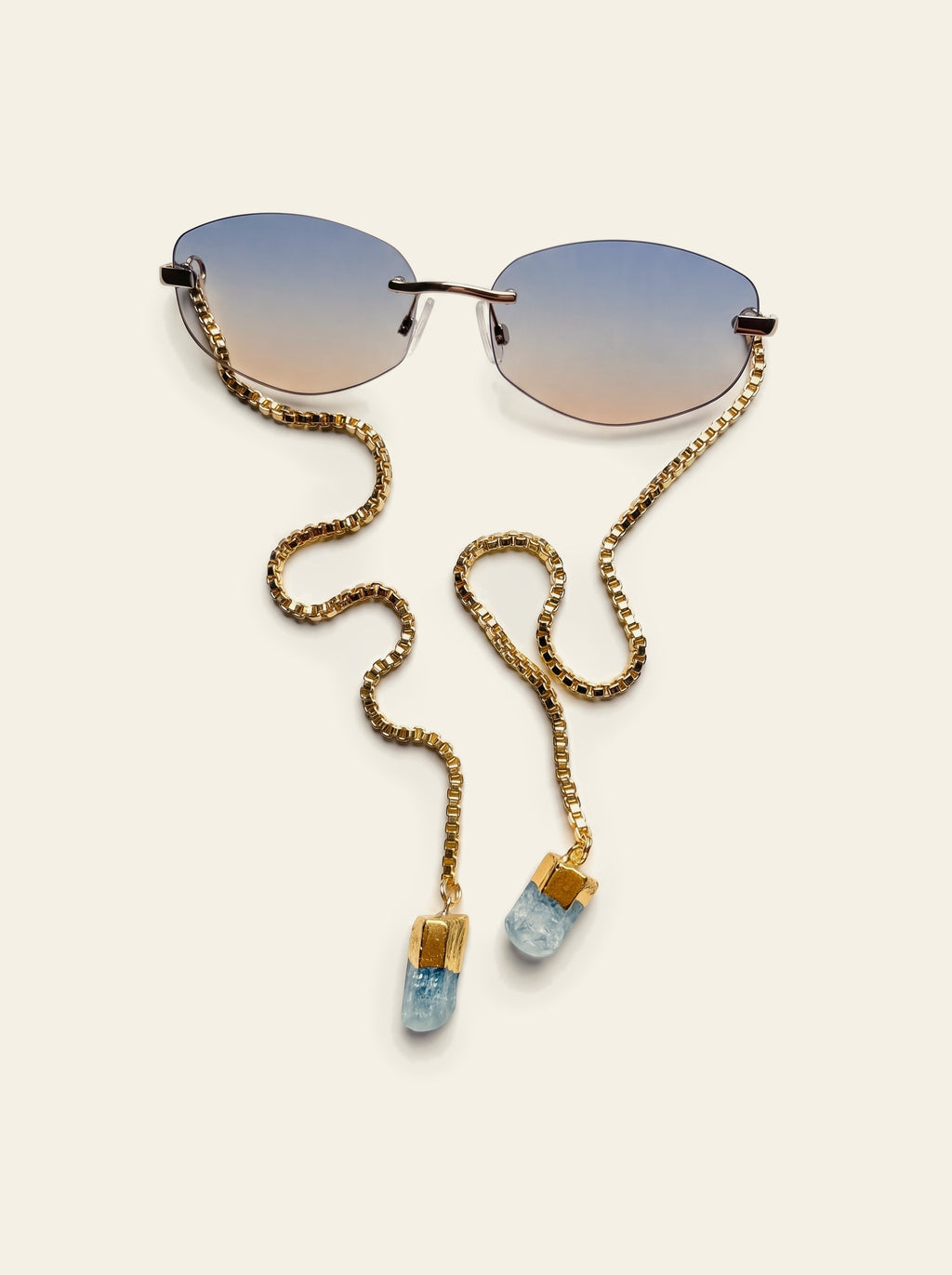 The Stresa - top view of rimless oval sunglasses with blue-grey gradient lenses and gold chain coiled beside them, blue quartz stones at each end