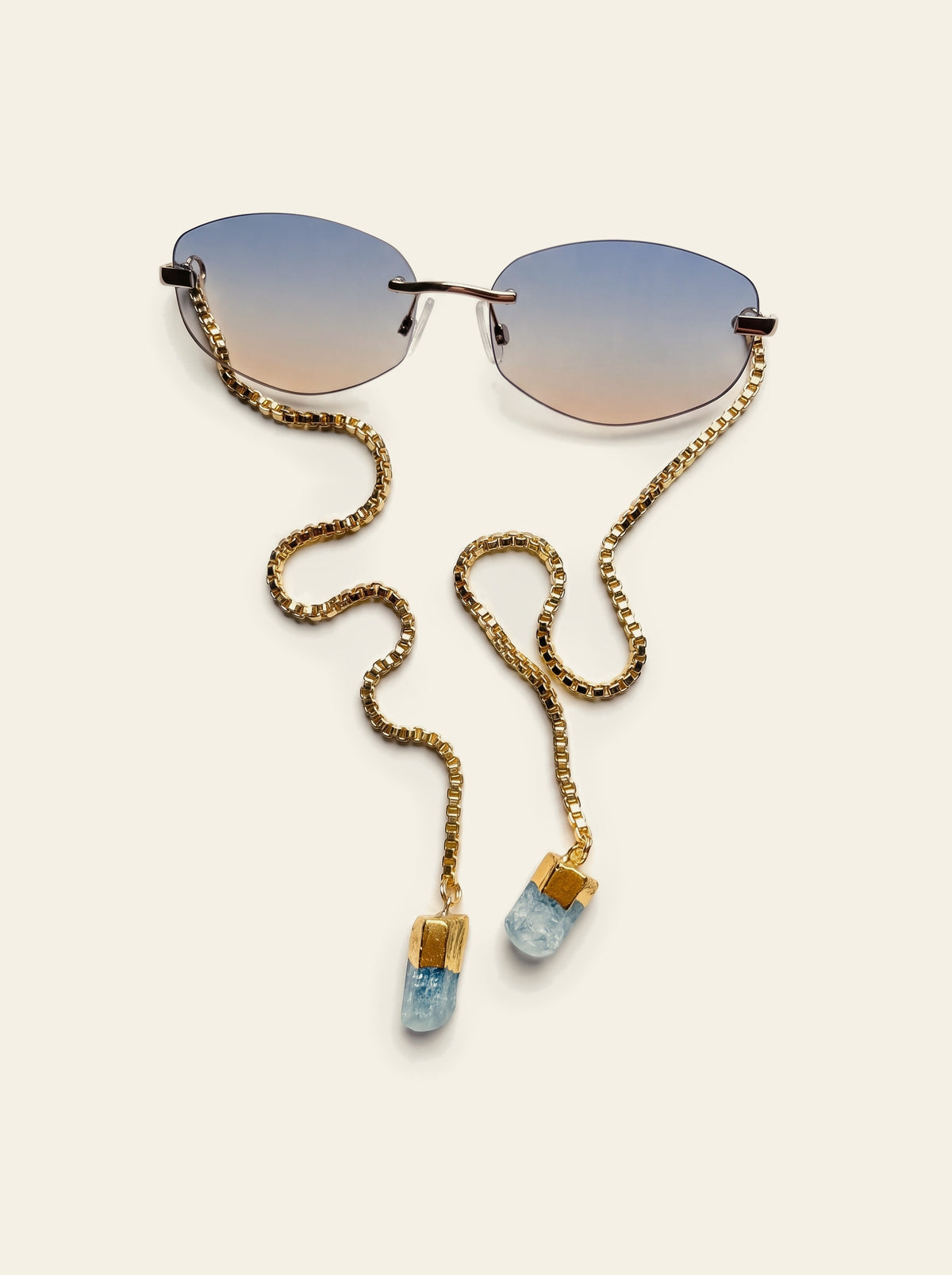 The Stresa - top view of rimless oval sunglasses with blue-grey gradient lenses and gold chain coiled beside them, blue quartz stones at each end
