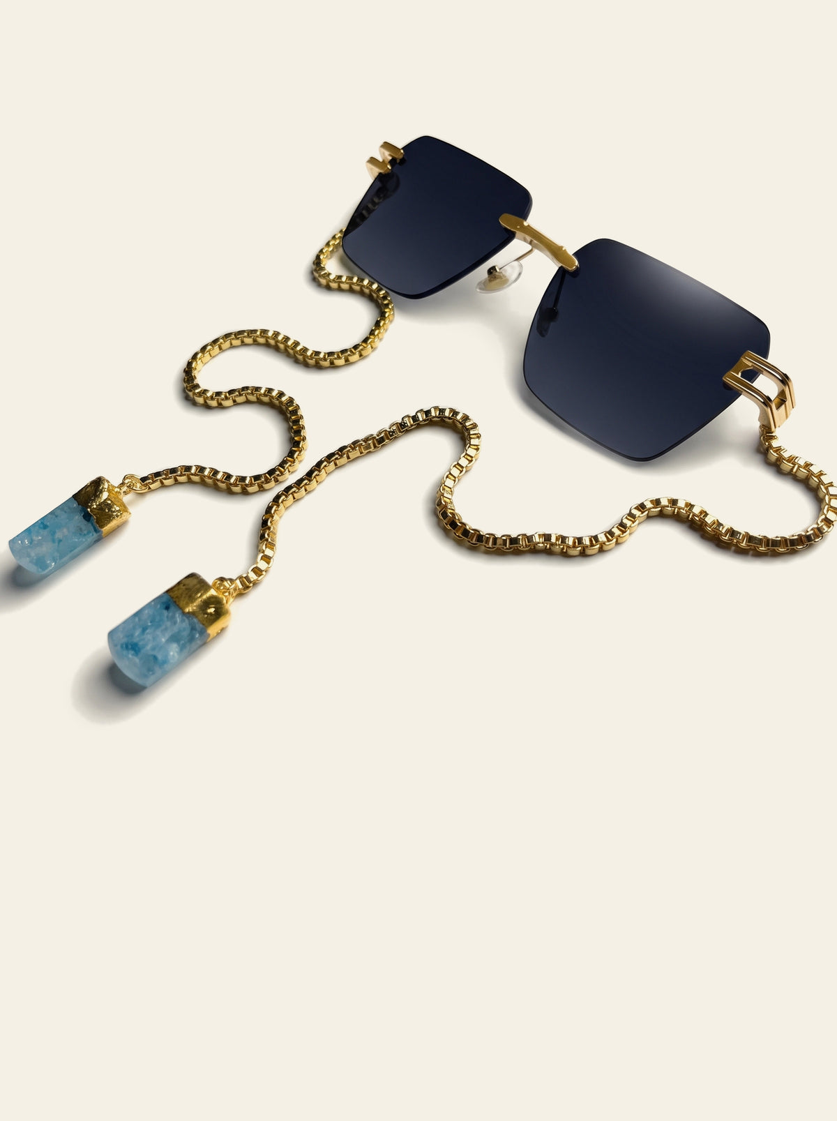 The Sardinia - rimless square gold sunglasses with deep ink-blue lenses, three-quarter view, gold chain and two pale blue quartz stones